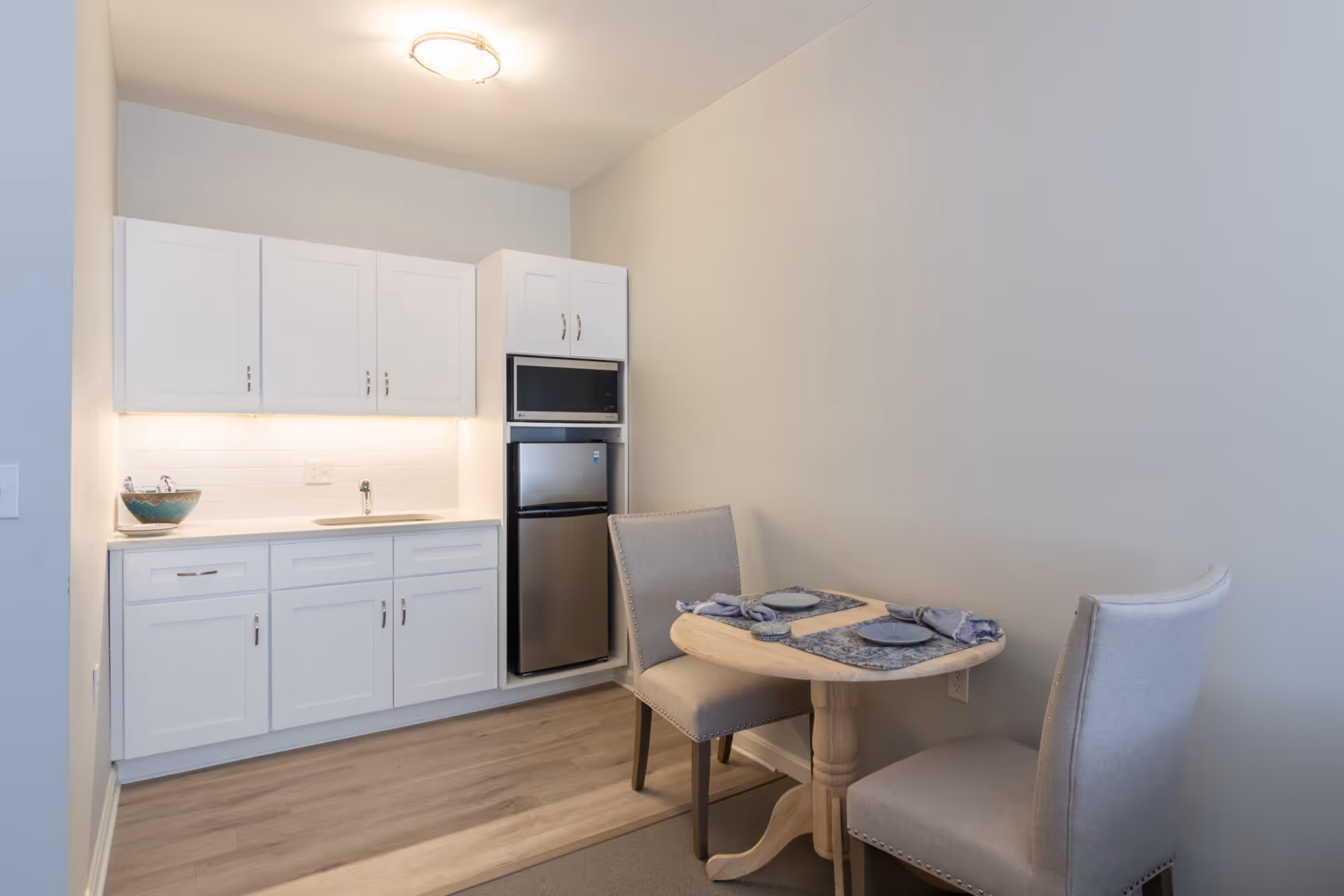 Small kitchenette with white cabinets, a mini fridge and microwave, and a small round dining table set for two.