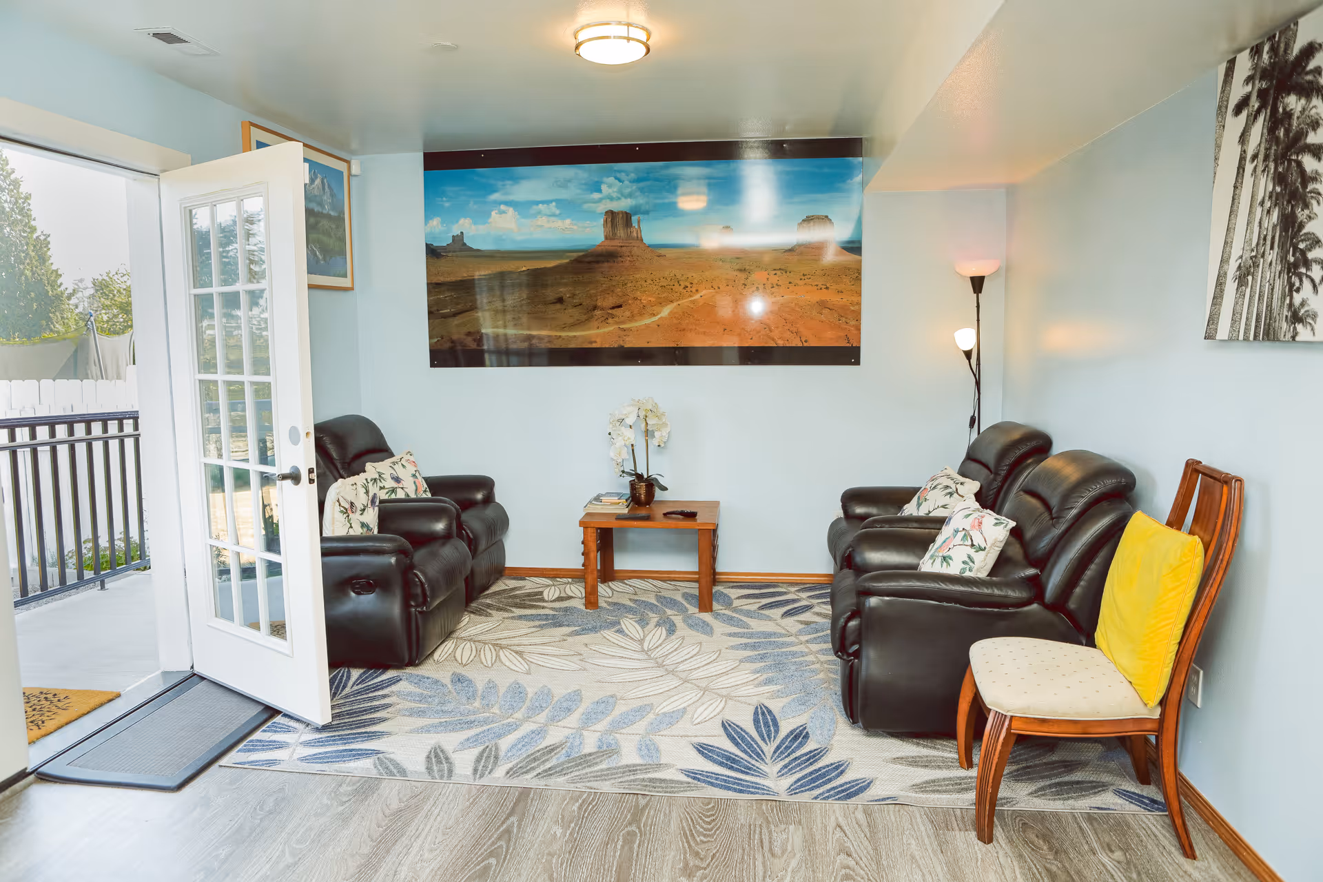 A cozy living room with light blue walls, featuring four black leather recliners with floral pillows arranged around a small wooden coffee table. A patterned area rug with leaf designs covers the floor. A wooden chair with a yellow cushion is placed on the right side. A large landscape photograph of a desert scene hangs on the wall. A floor lamp with two lights stands in the corner. A glass door on the left side opens to an outdoor area with a white fence and greenery.