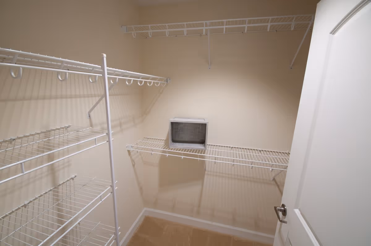 Empty walk-in closet with beige walls and carpeted floor, featuring white wire shelving units on three walls and a white door partially open.