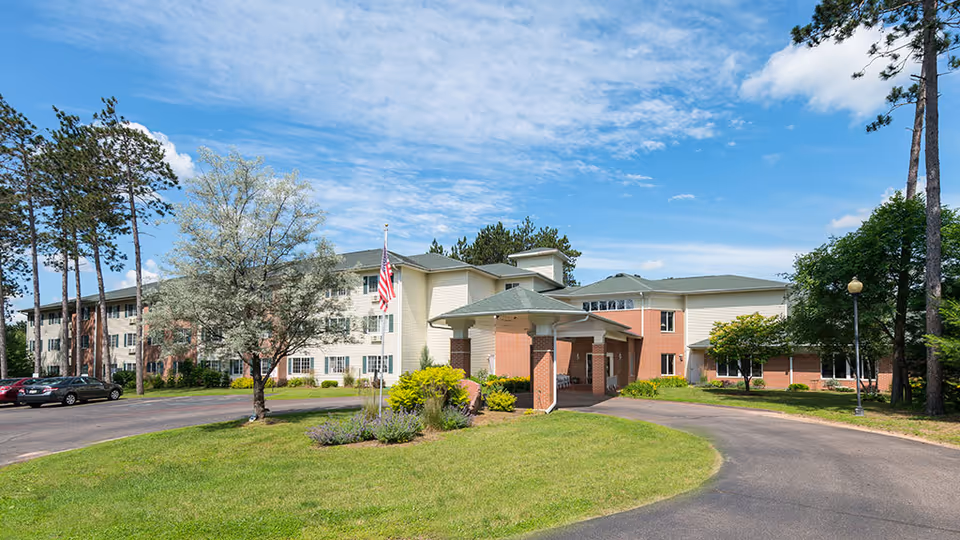 Exterior view of Park Vista Waupaca, a multi-story senior living facility with a covered entrance driveway, surrounded by green lawns, trees, and a clear blue sky.