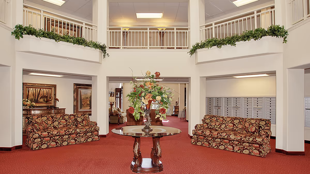 Interior view of a senior living facility lobby with two floral patterned sofas facing each other on a red carpet. A round wooden table with a large floral arrangement is centered in the foreground. The space features white walls, a second-floor balcony with white railings decorated with green plants, and mailboxes on the right side wall.