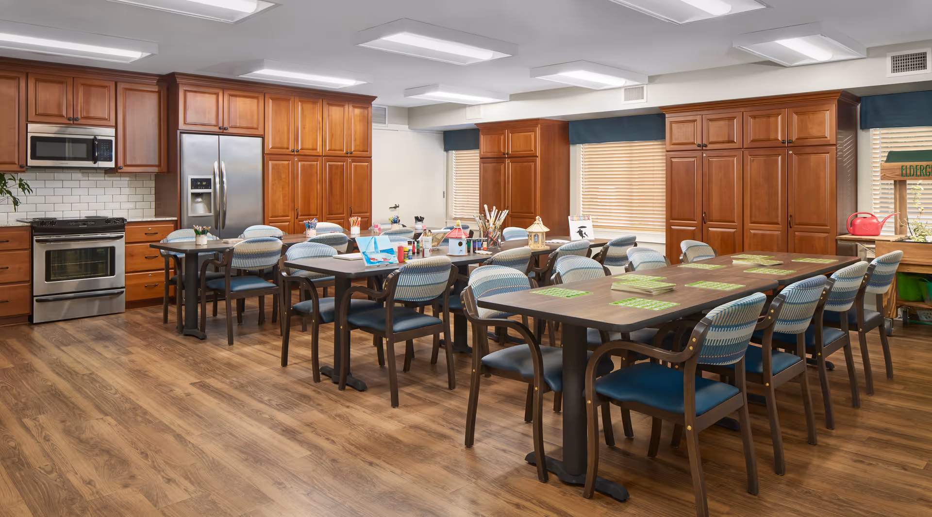 Communal dining and activity room with long tables and chairs, craft supplies on the tables, and a full kitchen with wood cabinets and stainless appliances.