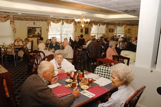 A dining room in a senior living facility with elderly residents seated at tables enjoying their meals. The room is decorated with patterned curtains, framed artwork, and chandeliers. Tables are set with red placemats, glasses of iced tea, and condiments.