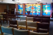 Rows of wooden chairs facing a colorful stained-glass window in a chapel or meeting room interior.