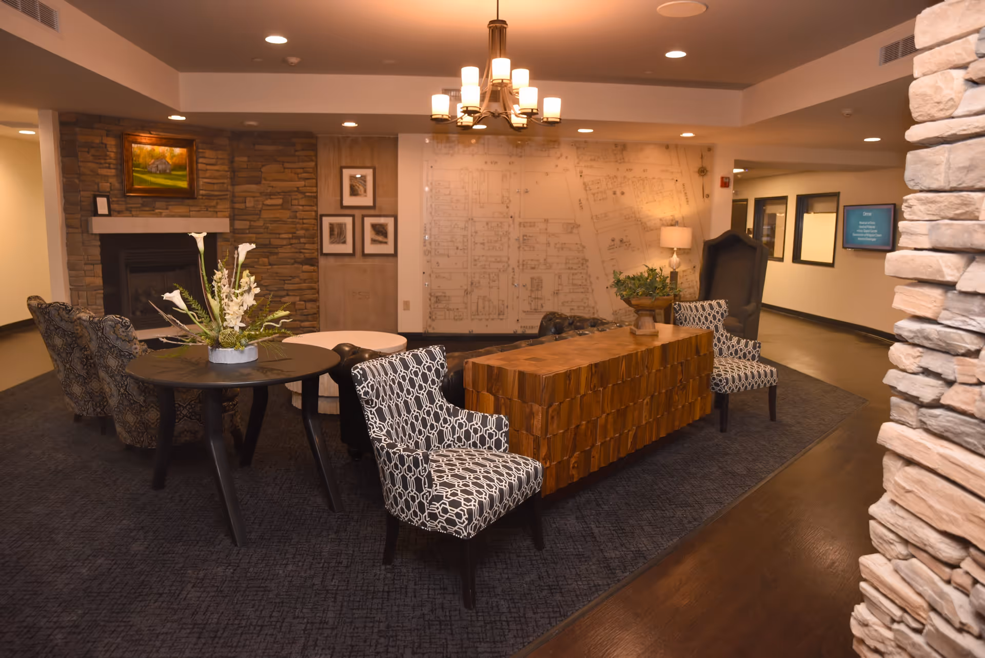 A cozy interior common area with patterned armchairs, a wooden block-style table, a round black table with a floral arrangement, and a stone fireplace with a painting above it. The walls feature framed pictures and a large architectural floor plan. The space is warmly lit with ceiling lights and a chandelier.