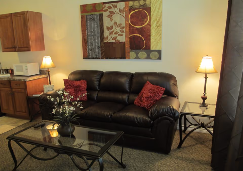 A cozy living room area featuring a dark brown leather sofa with two red decorative pillows. In front of the sofa is a glass coffee table with a vase of white flowers. To the right of the sofa is a glass side table with a lamp, and to the left is a small kitchen area with wooden cabinets, a microwave, and a lamp. A colorful abstract painting hangs on the wall above the sofa.