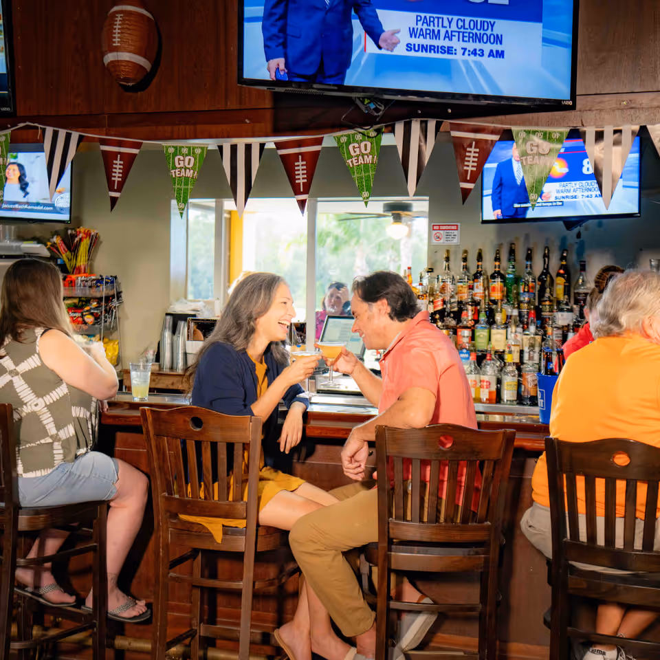A group of people sitting on wooden bar stools at a bar counter decorated with football-themed pennant banners. Two people in the center are smiling and clinking their cocktail glasses. Behind the bar, there is a large selection of liquor bottles and multiple televisions mounted on the wall showing a weather forecast.