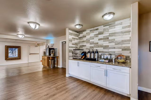 Interior view of a senior living facility hallway with a coffee and snack station on the right featuring white cabinets, a granite countertop, and a tiled backsplash. The hallway has wood flooring, beige walls, ceiling lights, and a framed picture on the far wall.
