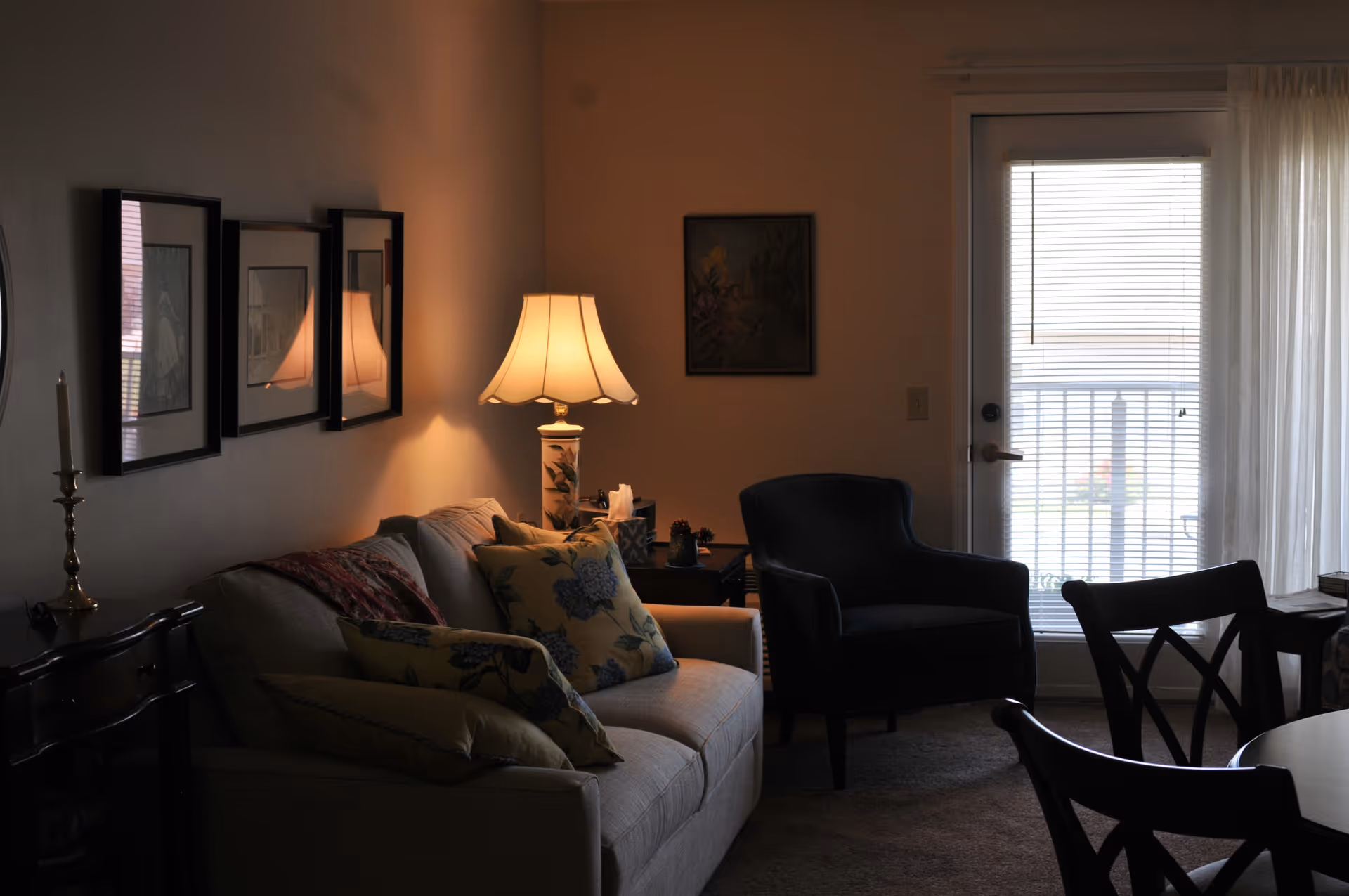 A cozy living room with a beige sofa adorned with floral cushions, a lit table lamp on a side table, a dark armchair, framed pictures on the wall, and a glass door with blinds leading outside.