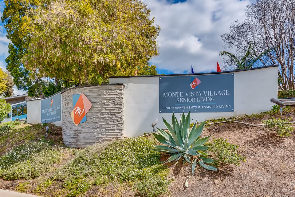 Outdoor view of the entrance sign for Monte Vista Village Senior Living, featuring a stone and white wall with the facility's logo and name, surrounded by greenery and plants under a partly cloudy sky.
