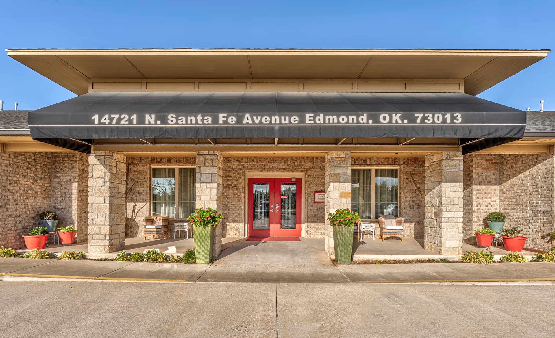 Front exterior view of a senior living facility with a black awning displaying the address 14721 N. Santa Fe Avenue Edmond, OK 73013. The building has stone pillars, red double doors, and potted plants on either side of the entrance.