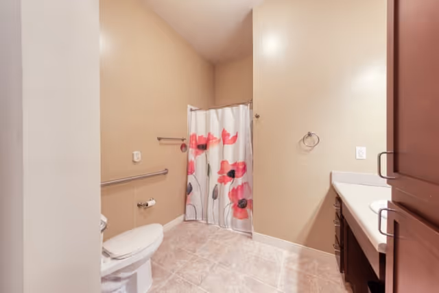 A bathroom with beige walls and tiled floor featuring a white toilet with a grab bar beside it, a shower area with a curtain decorated with large red and pink flowers, and a vanity with a sink and dark wood cabinets.