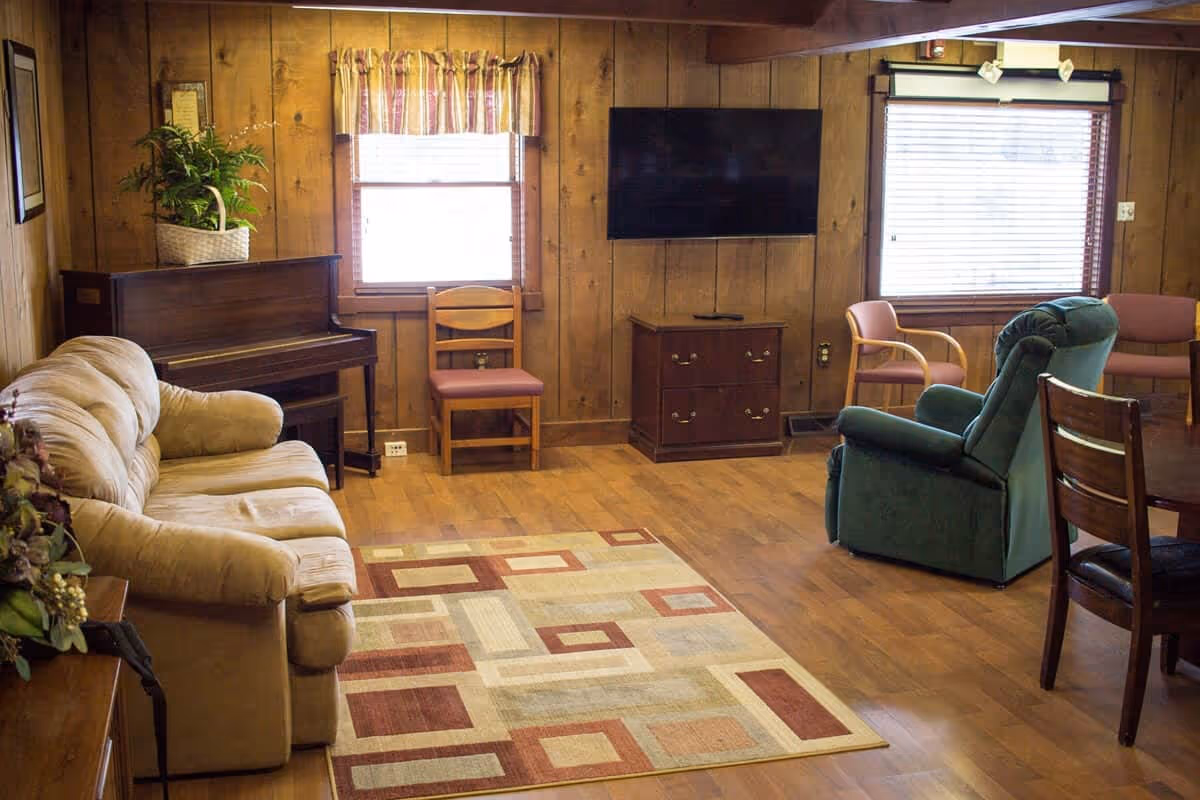 Cozy wood-paneled common living room with a sofa, recliner, TV, piano, chairs, and area rug.