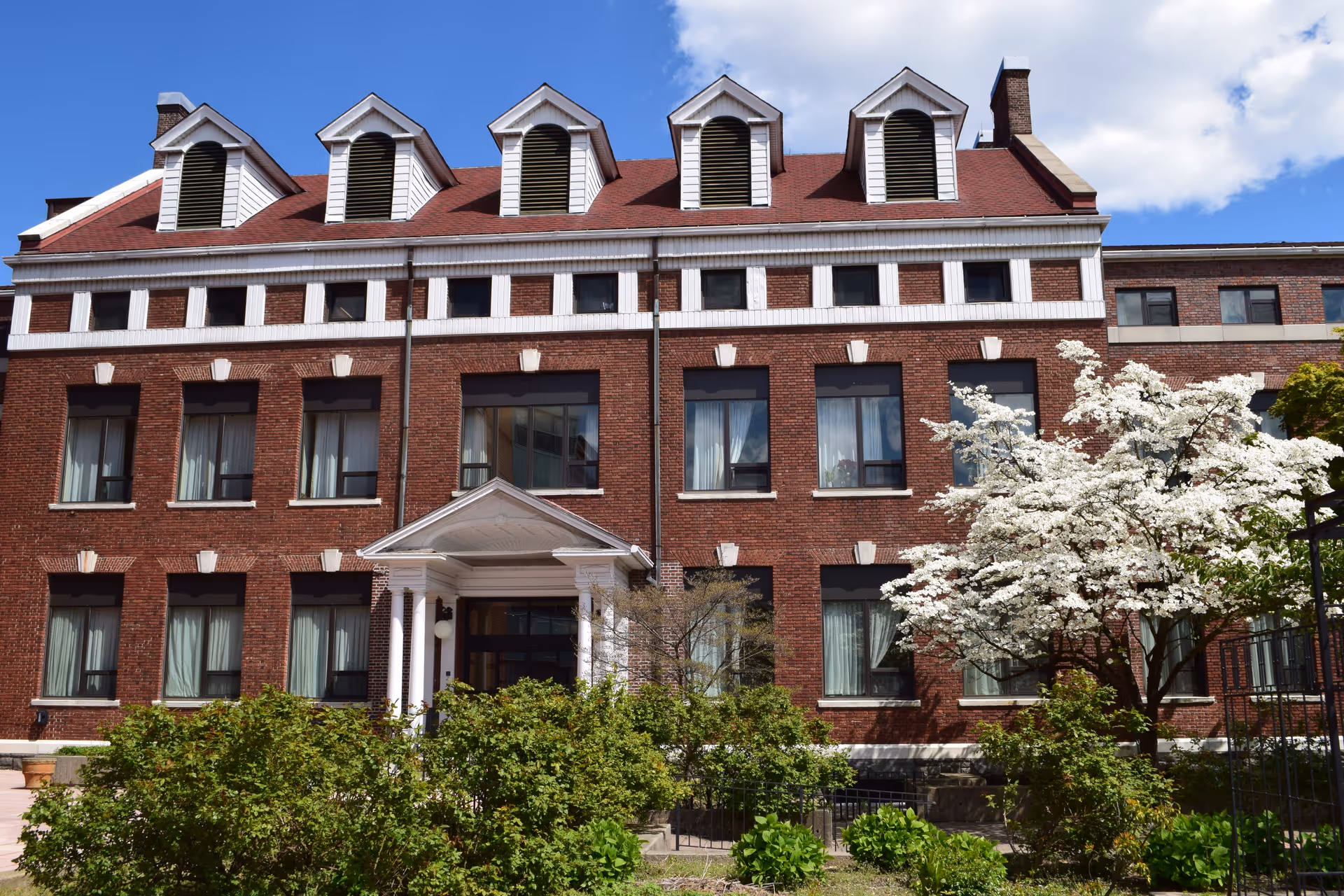 Front view of a three-story red brick assisted living building with white-trimmed dormer windows, a columned entrance, and flowering trees and shrubs.