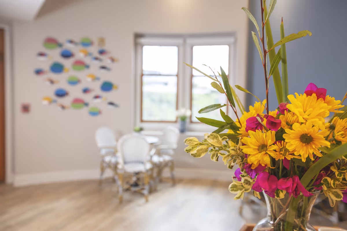 Vase of bright yellow and pink flowers in the foreground with a blurred communal dining area and windows in the background.