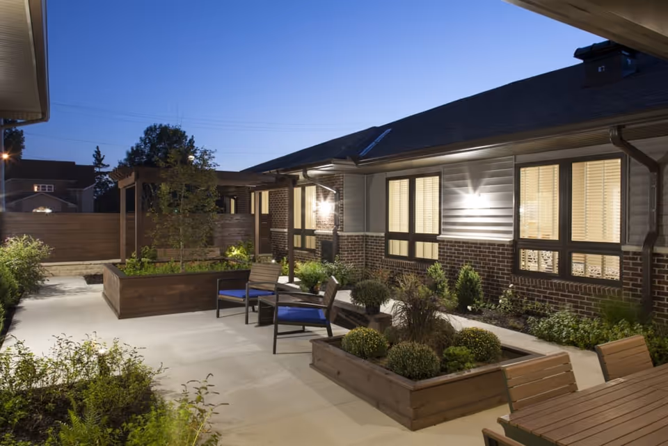 Outdoor patio area at dusk with raised wooden garden beds containing plants and shrubs, several chairs with blue cushions, a wooden pergola, and a building with lit windows in the background.