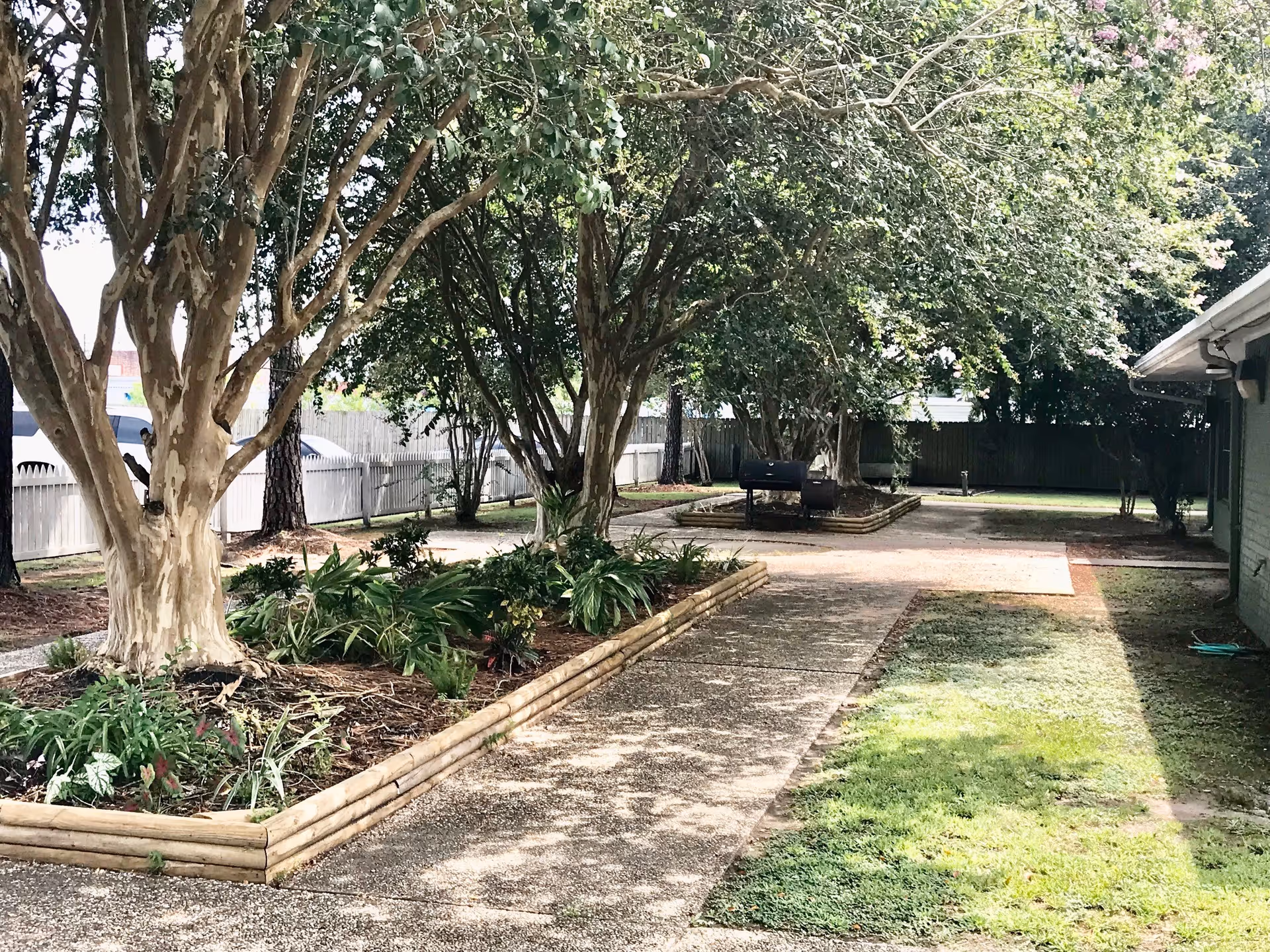 Outdoor garden area at Oak Grove Nursing Home with a paved walkway, raised flower beds with trees and plants, a barbecue grill, and a wooden fence in the background.