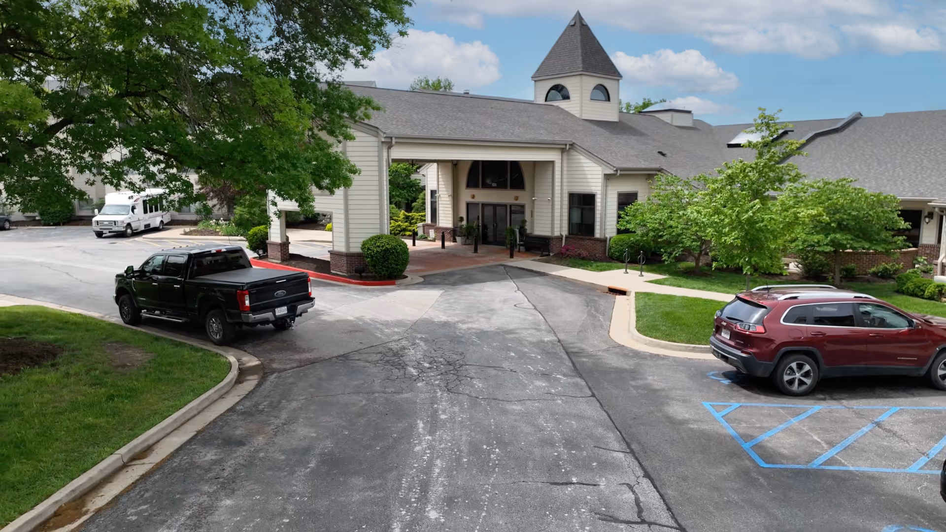 Front exterior view of a senior living facility named Unbridled Living of Columbia, showing a covered entrance with a peaked roof and a small tower. There are two vehicles parked near the entrance, surrounded by green trees and landscaping under a partly cloudy sky.