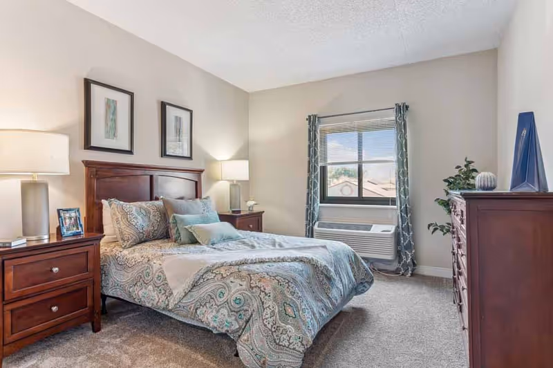 A neatly made bed with patterned bedding in a bedroom featuring wooden furniture including two nightstands with lamps, a dresser, and a window with patterned curtains and a wall-mounted air conditioning unit.