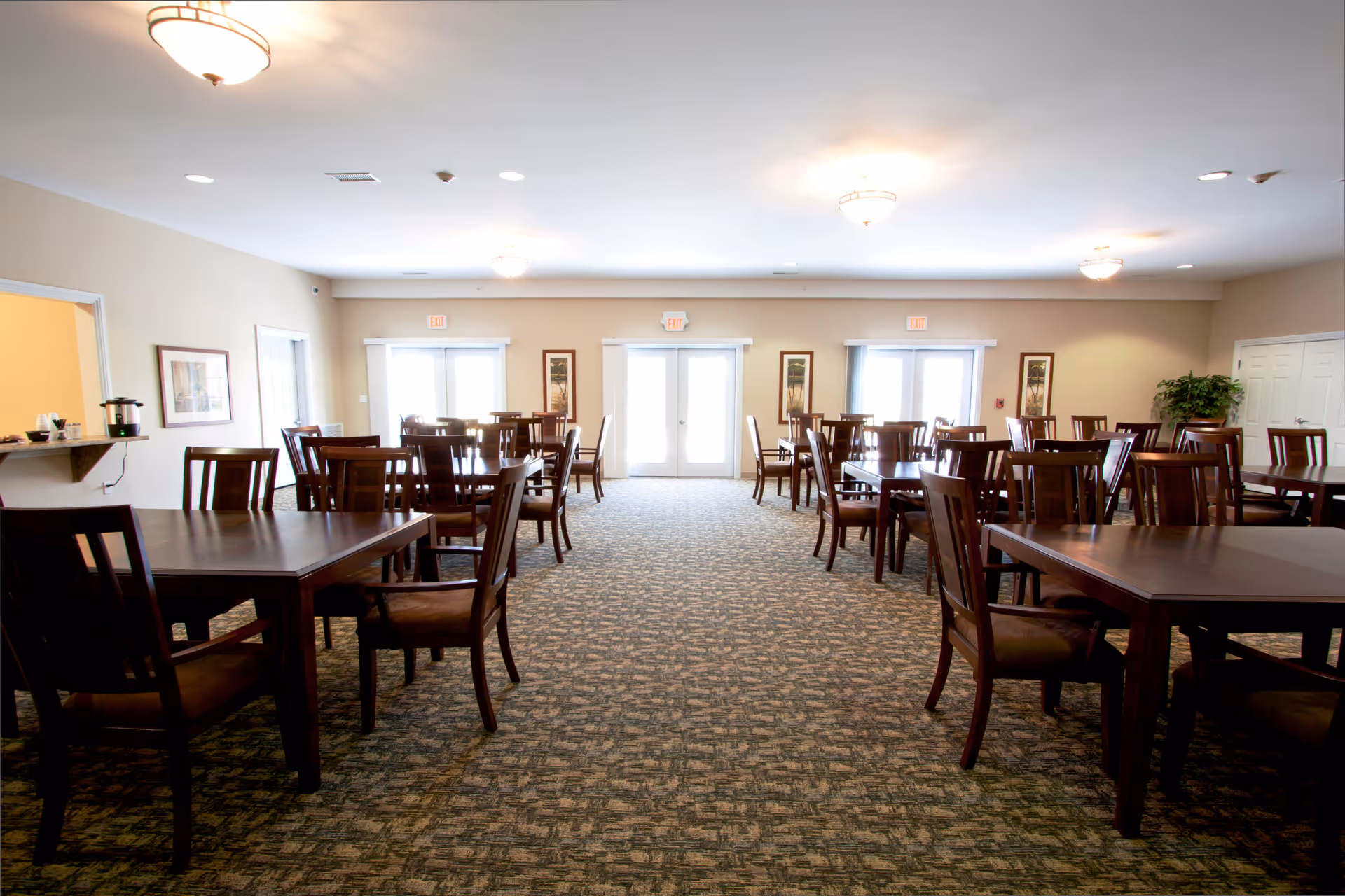 A spacious dining room with multiple dark wood tables and chairs arranged neatly on a patterned carpet. The room is well-lit with ceiling lights and has beige walls adorned with framed artwork. Large windows and glass doors at the far end allow natural light to enter the room.