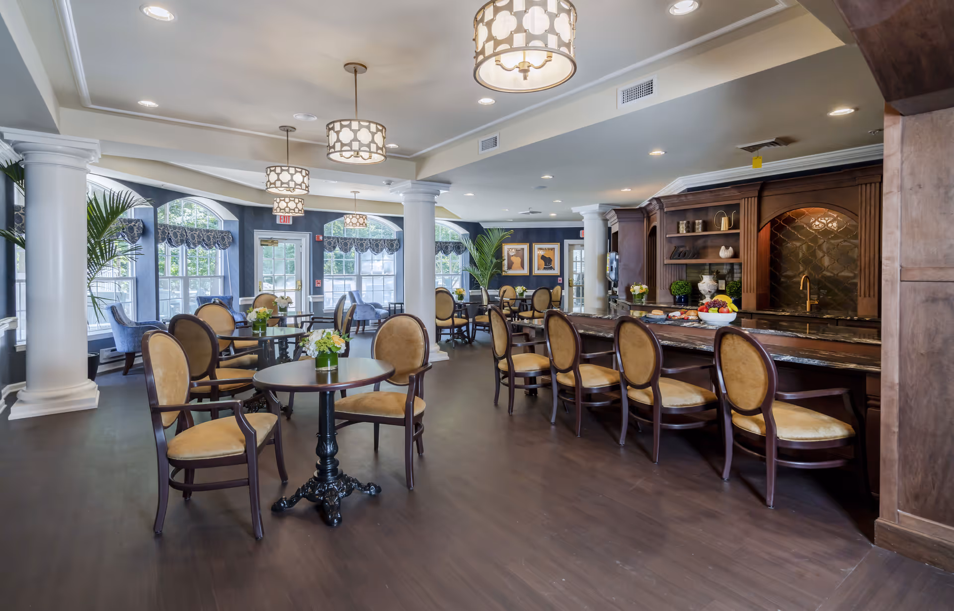 A spacious and elegant dining area with round tables and cushioned chairs arranged neatly. The room features large windows with decorative valances, white columns, and hanging pendant lights. There is a dark wood bar counter with high chairs on the right side, and the overall decor is warm and inviting with plants and framed artwork on the walls.