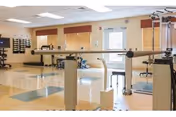 Bright rehabilitation therapy room with parallel walking bars, exercise equipment, stools and polished tile floor.