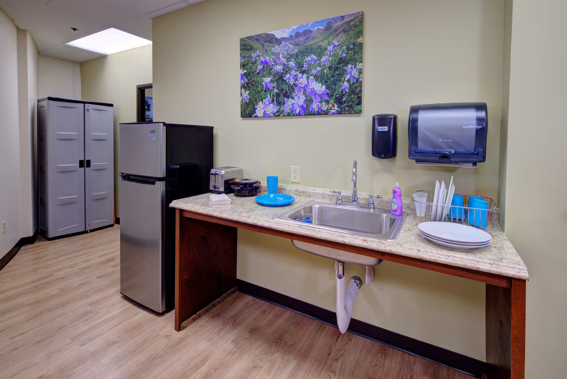 A small kitchenette area with a stainless steel refrigerator, a countertop with a sink, a toaster, a plate with a blue cup, and a dish rack holding plates and blue cups. There is a soap dispenser and a paper towel dispenser mounted on the wall above the sink. A colorful floral painting hangs on the wall above the countertop. The floor is light wood, and there are two tall storage cabinets in the background.
