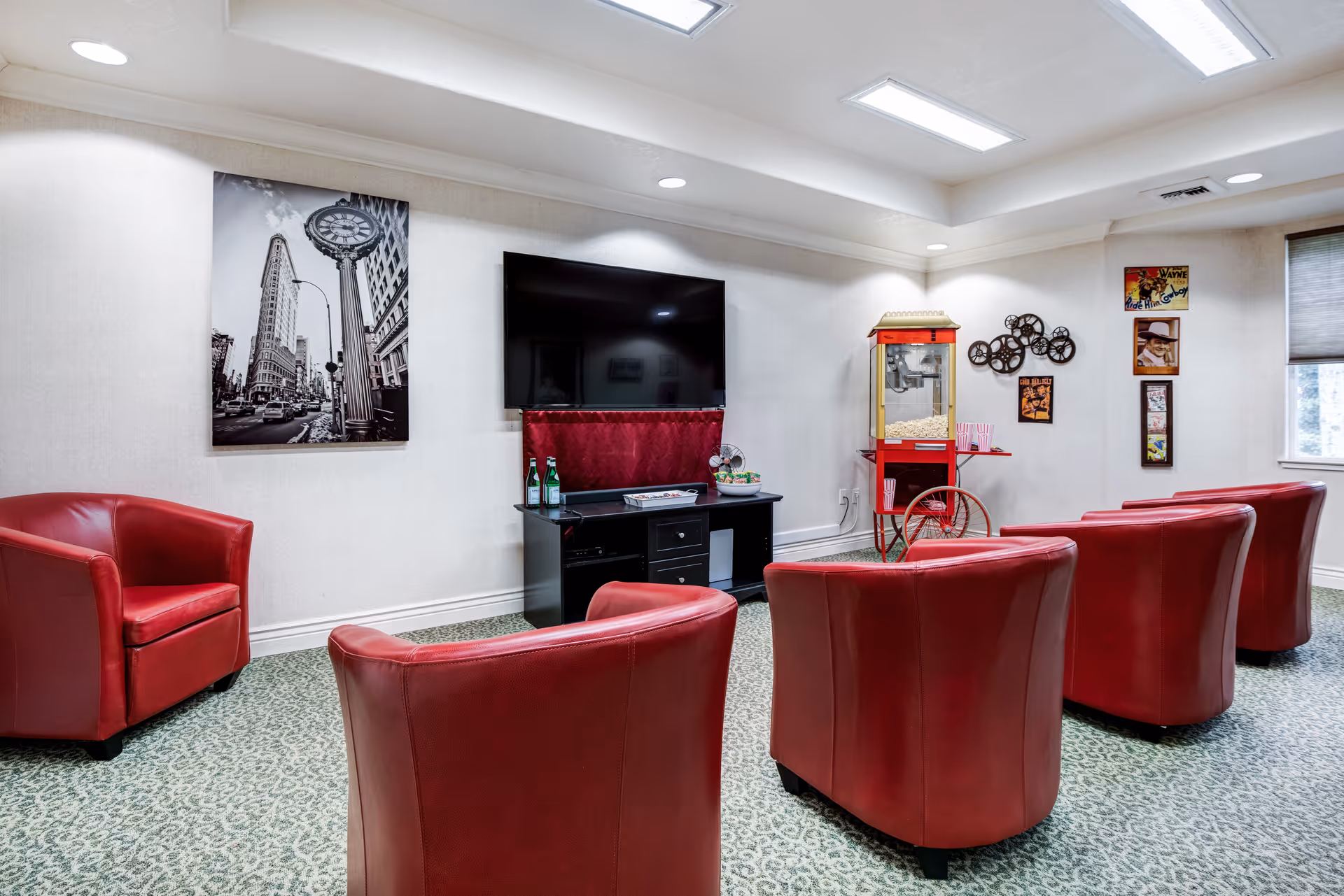 A cozy lounge area with five red leather armchairs arranged facing a flat-screen TV mounted on a wall. Below the TV is a black entertainment console with bottles and a decorative item. To the right, there is a vintage-style popcorn machine and wall decorations including film reels and framed pictures. The room has patterned carpet flooring and a window with blinds on the right side.