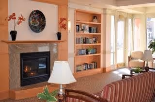 A cozy living room with a fireplace surrounded by a wooden mantel and two vases with orange flowers on top. To the right of the fireplace is a built-in bookshelf filled with books and decorative items. The room has large windows allowing natural light to fill the space. There is a striped sofa in the foreground, a table lamp, and a comfortable armchair near the windows.