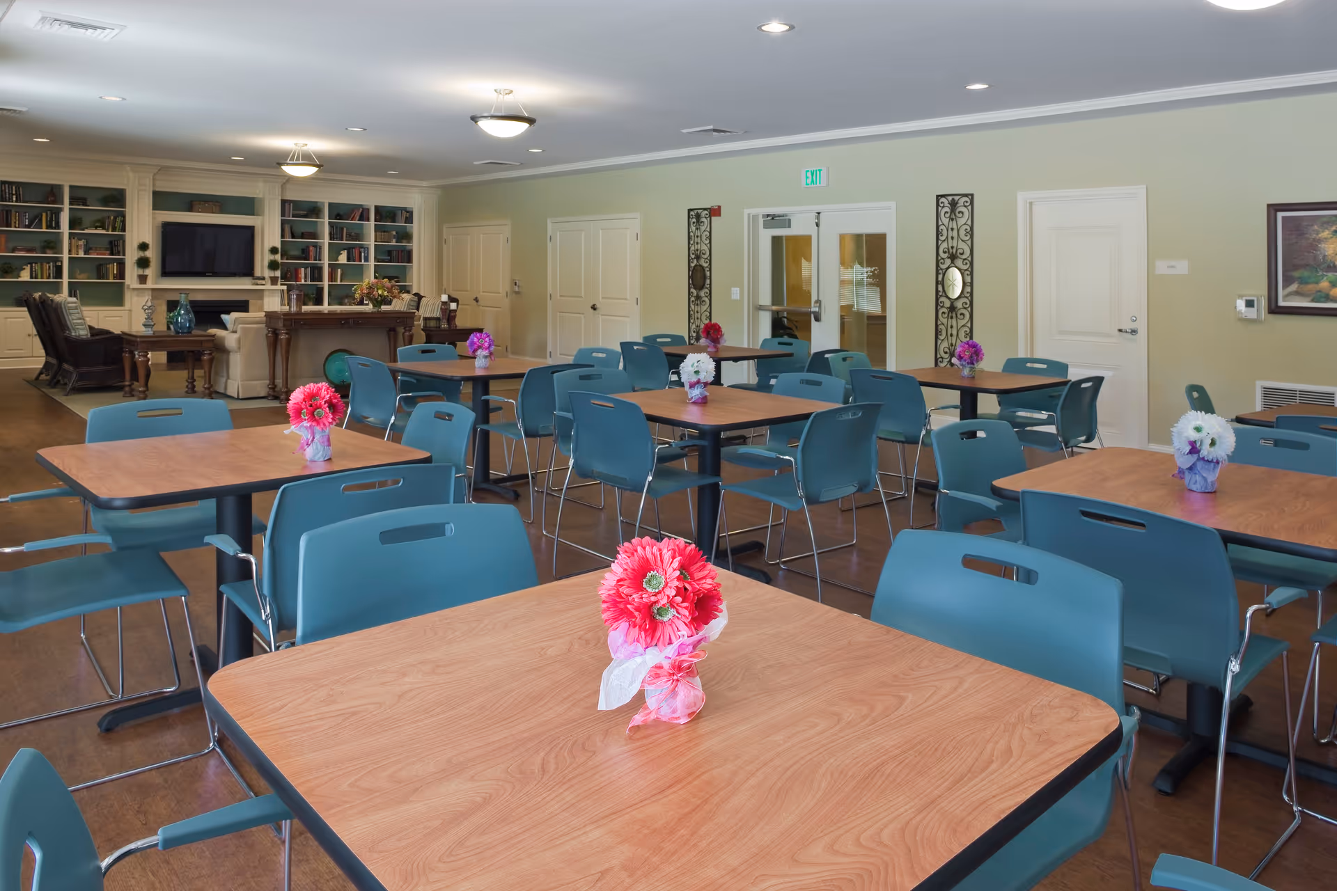 A spacious dining room with multiple wooden tables and teal chairs arranged neatly. Each table has a small floral centerpiece with colorful flowers. In the background, there is a cozy living area with bookshelves, a television, and comfortable seating. The walls are painted light green, and the room is well-lit with ceiling lights.