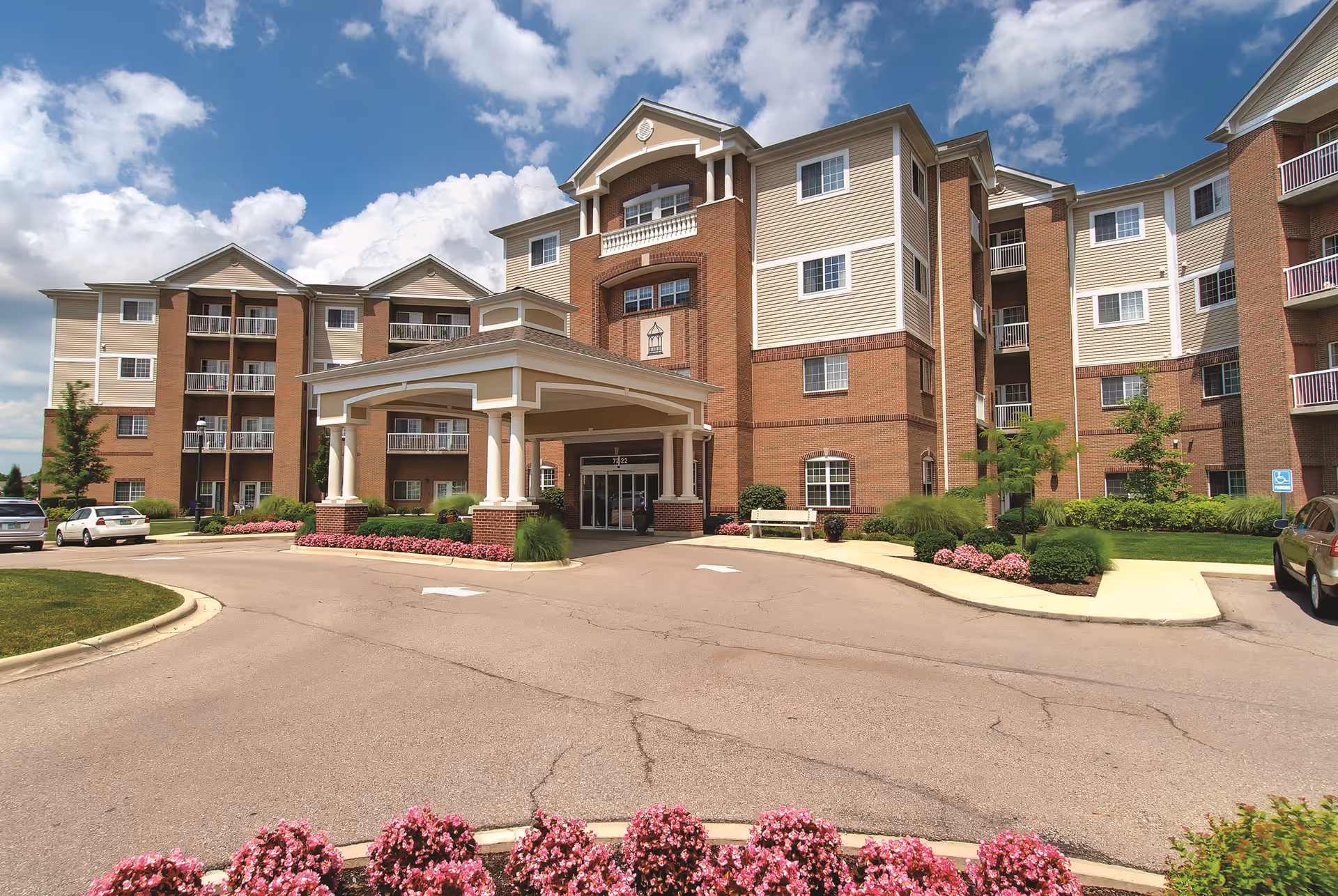 Exterior view of a multi-story senior living facility building with a covered entrance driveway, landscaped flower beds, and several parked cars under a partly cloudy sky.