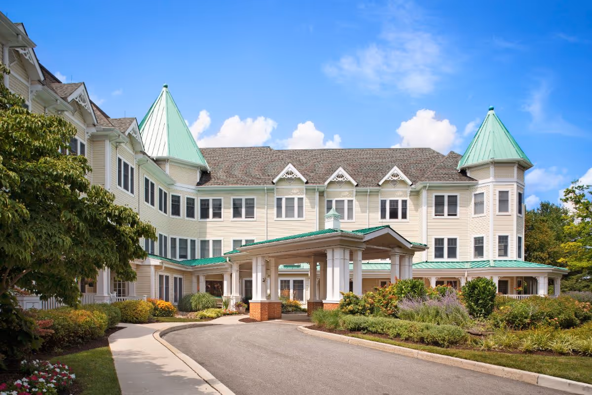 Front exterior view of Morningside House of Blue Bell, a large cream-colored building with green roofs and two prominent turret-style towers. The entrance features a covered driveway with white columns and brick bases, surrounded by landscaped gardens and a clear blue sky with some clouds.