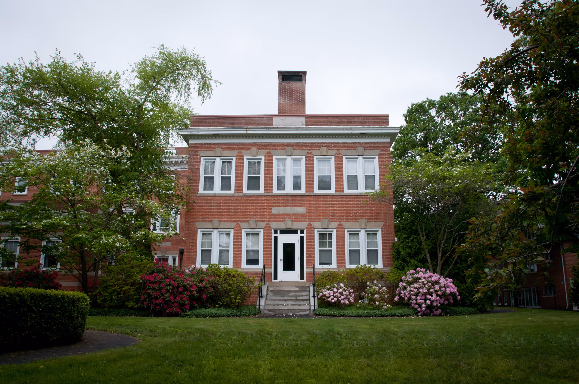 Front exterior view of a two-story red brick building with white-framed windows and a central door. The building is surrounded by green trees, bushes, and flowering plants, with a well-maintained lawn in the foreground under an overcast sky.