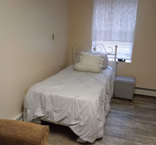 A small bedroom with a single bed covered in a white comforter and two decorative pillows. The bed has a white metal headboard. There is a window with blinds partially closed behind the bed, and a small gray ottoman next to the bed. The floor is wood-style laminate, and a brown upholstered chair is partially visible in the foreground.