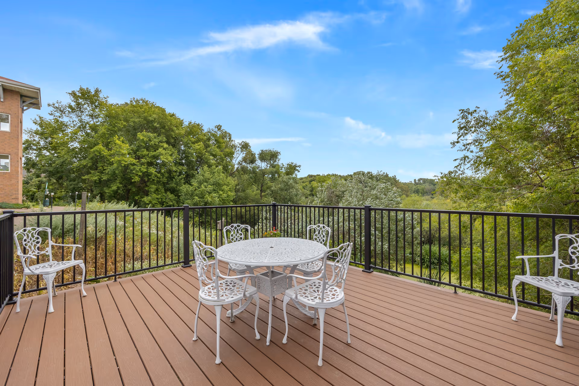 Outdoor deck with a white metal table and chairs overlooking trees and greenery under a blue sky.