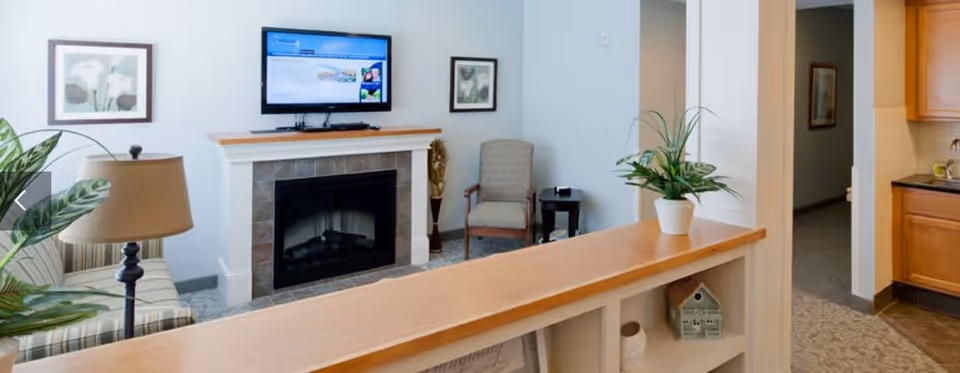 A cozy living room area with a fireplace and a mounted flat-screen TV above it. There is a striped sofa with a lamp on the left, a single armchair with a small side table on the right, and a wooden shelf with a potted plant in the foreground. The room has light-colored walls and carpeted flooring, with a partial view of a kitchen area to the right.