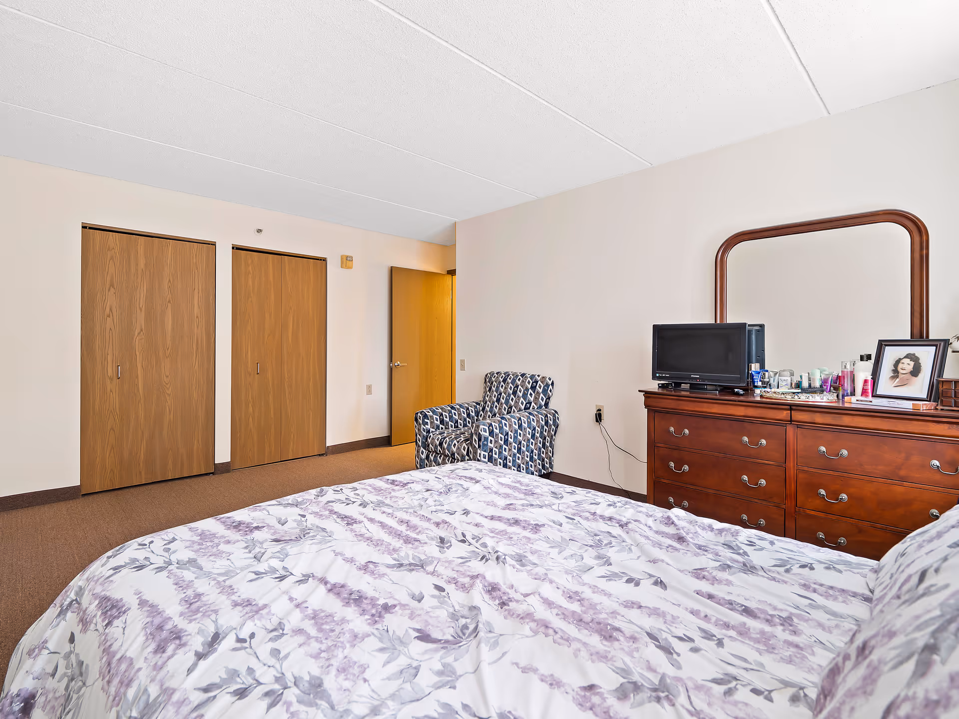 A bedroom with a bed covered in a white and purple floral comforter, a wooden dresser with a mirror, a small TV, and various personal items on top. There is a patterned armchair near two closed wooden closet doors and an open wooden door leading out of the room. The walls are light-colored and the floor is carpeted.