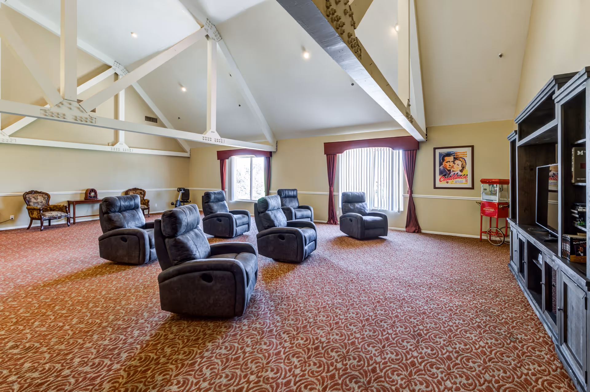 A spacious room with a high vaulted ceiling featuring exposed white beams. The room has six gray recliner chairs arranged in two rows facing a large wooden entertainment center with a TV. There is a vintage-style popcorn machine and a framed Casablanca movie poster on the wall. The floor is covered with a patterned red carpet, and there are two windows with red curtains allowing natural light into the room. Two upholstered armchairs and a small table are placed against the far wall.