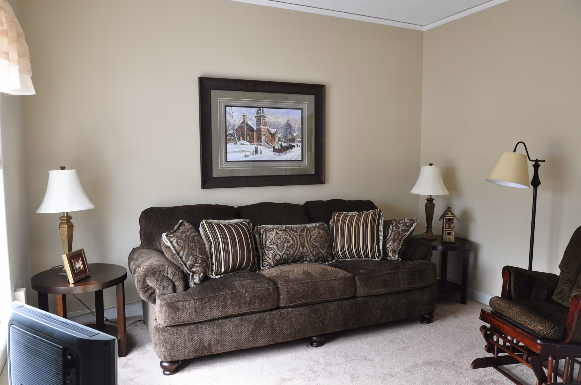 A cozy living room with a brown upholstered sofa adorned with patterned cushions, two side tables each holding a lamp, a framed winter scene painting on the beige wall, a wooden rocking chair with cushions, and a television partially visible in the foreground.