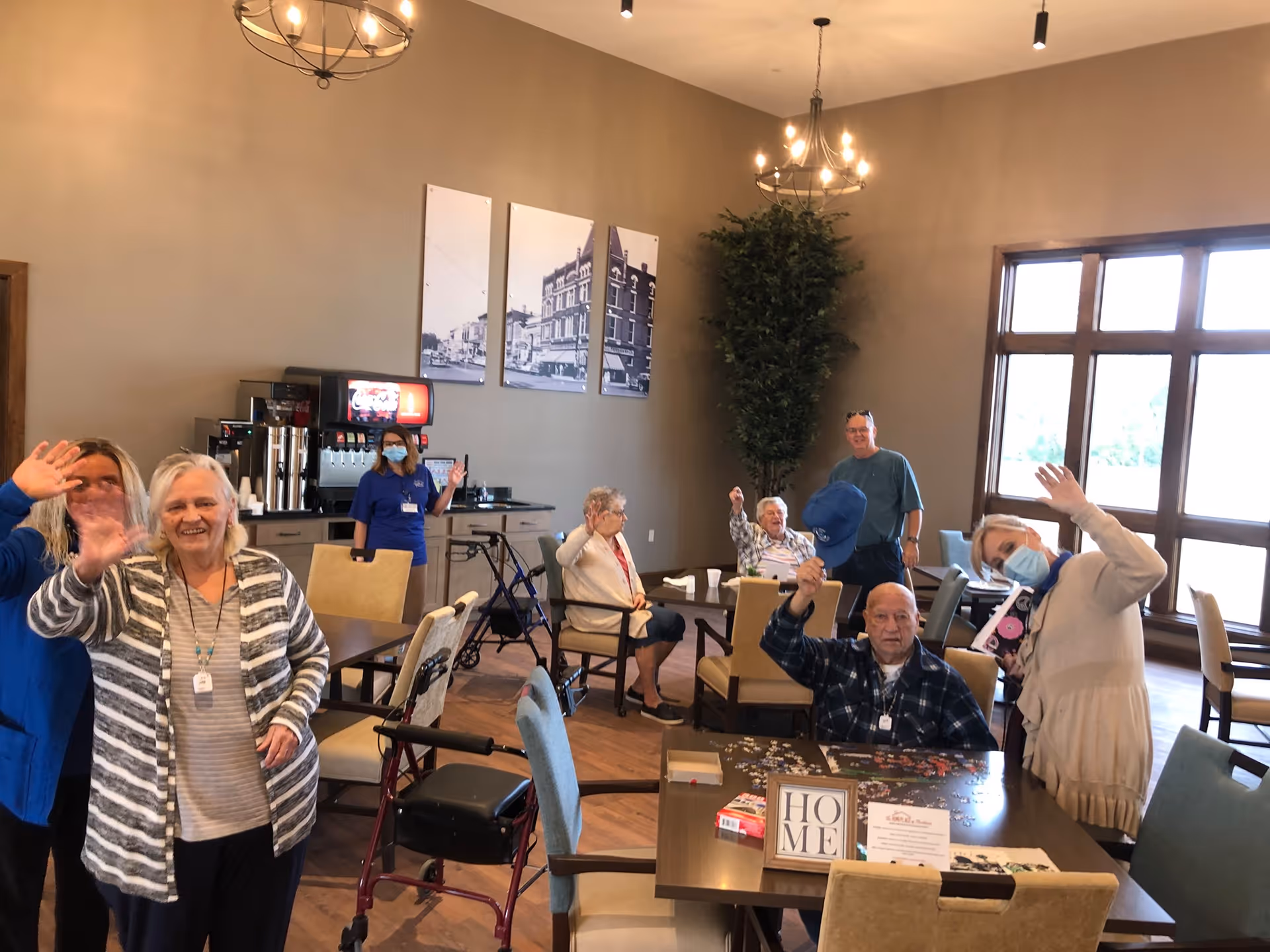 A group of elderly residents and staff in a common room of a senior living facility. Some residents are seated at tables working on a puzzle, while others are waving and smiling at the camera. The room has large windows, beige walls, chandeliers, and framed black-and-white photos on the wall.
