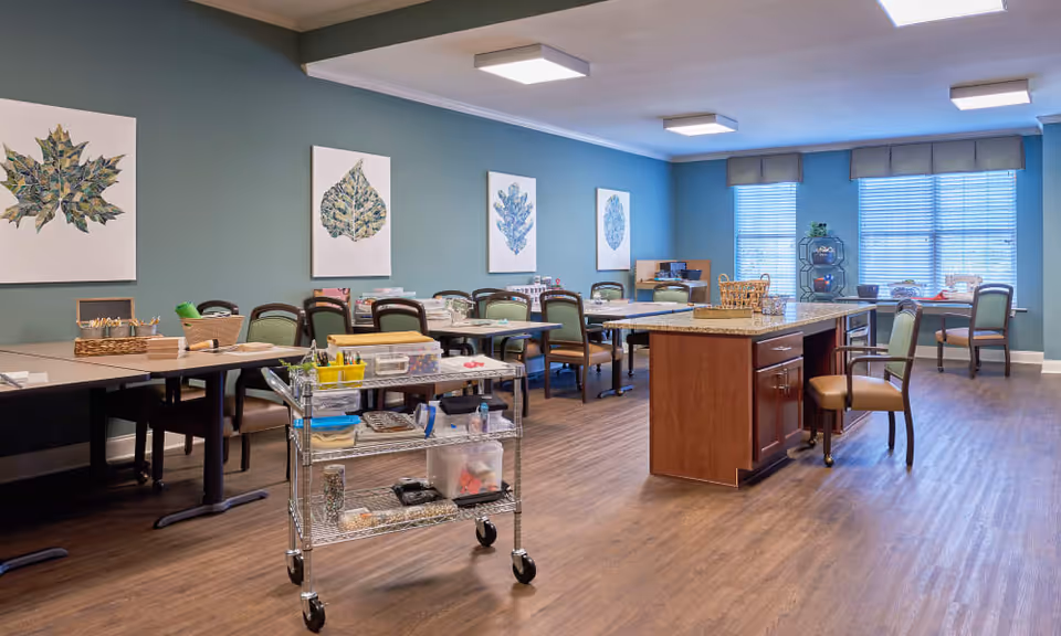 A spacious activity room with multiple tables and chairs arranged around the room. The walls are painted teal and decorated with framed leaf artwork. There is a large island with a granite countertop in the center, and a metal cart with various craft supplies. Large windows with blinds allow natural light to fill the room.