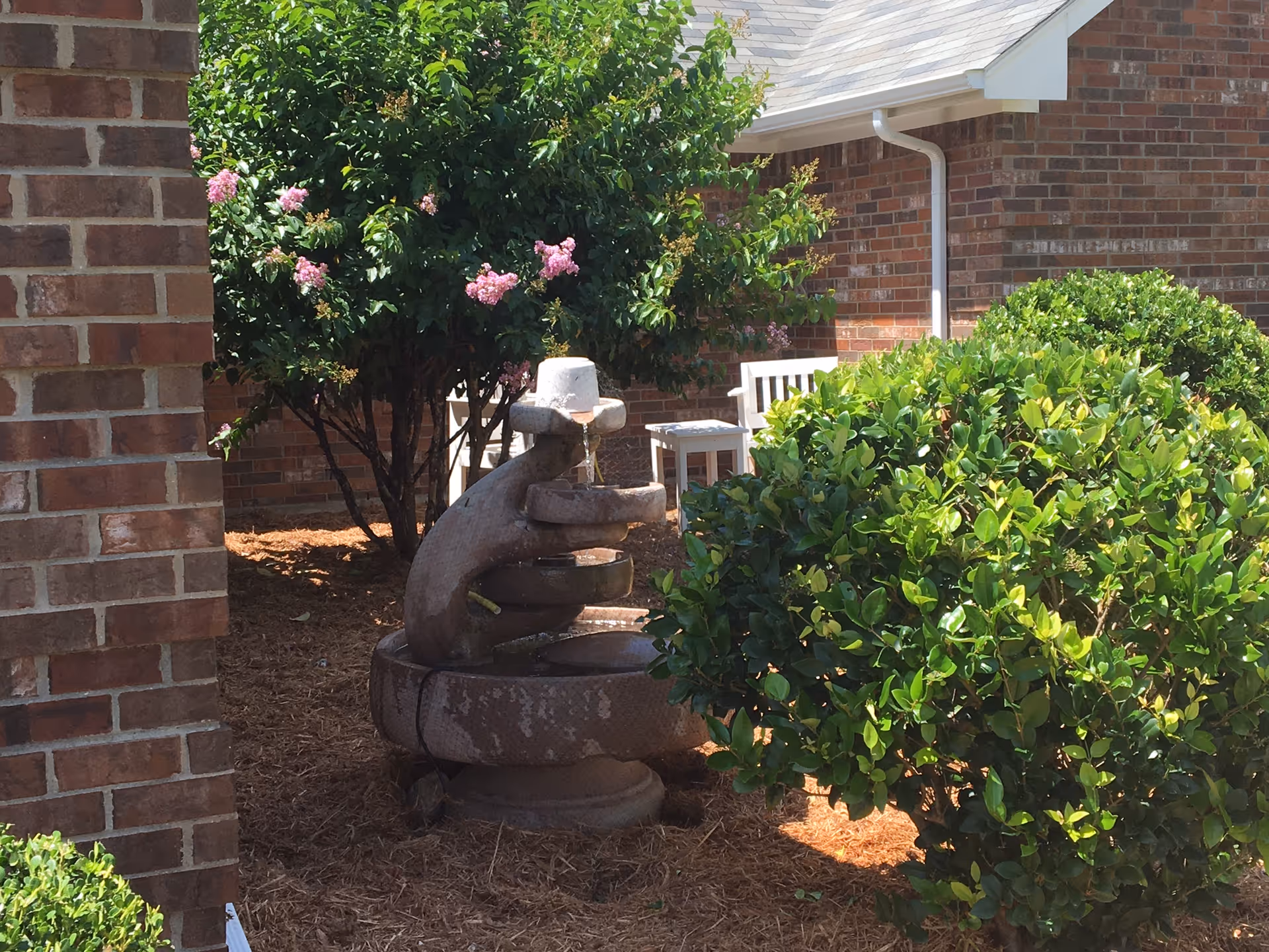 Outdoor garden area with a multi-tiered stone water fountain surrounded by green bushes and a flowering tree with pink blossoms. A brick building and white outdoor chairs are visible in the background.