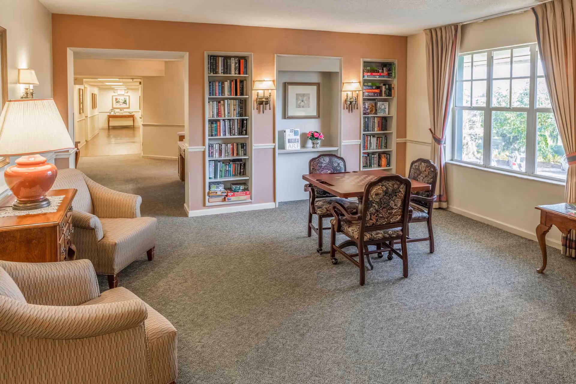 Bright common lounge with upholstered chairs, a card table and chairs, built-in bookshelves, and a large window.