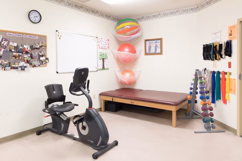 A therapy room with exercise equipment including a recumbent stationary bike, a padded therapy table, colorful exercise balls stored in wall-mounted nets, a rack of small dumbbells, and various resistance bands hanging on the wall. A bulletin board with photos and a whiteboard are also visible on the walls.