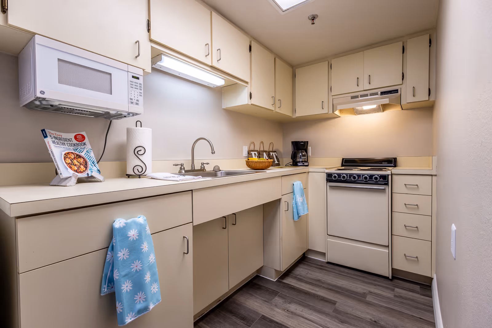 A compact kitchen with beige cabinets and drawers, a white microwave mounted above the counter, a stainless steel sink with a faucet, a stove with an overhead vent, a coffee maker, and a cookbook displayed on a stand. Two blue towels with white flower patterns hang from the cabinet handles. The floor has dark wood-style flooring.