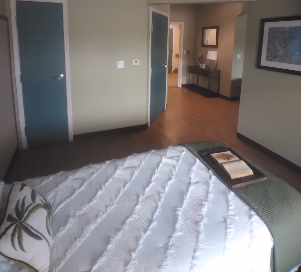 A neatly made bed with a tray and book in the foreground of a bedroom showing green doors, hardwood floors, and a hallway with a console table.