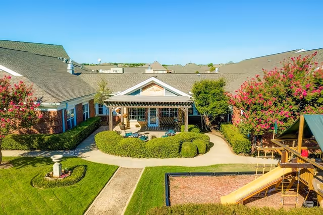 Outdoor courtyard area of a senior living facility with a covered patio featuring seating and a pergola, surrounded by manicured hedges and trees with pink flowers. There is a playground with a slide and climbing structure on the right side, and a birdbath on the left side, all set against a backdrop of single-story brick buildings with green roofs.