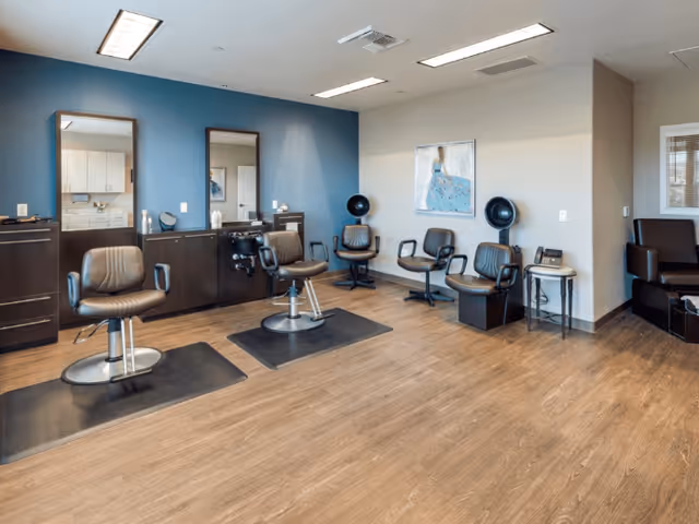 Interior view of a salon area with two styling chairs in front of mirrors mounted on a blue accent wall, additional seating with hair drying stations, and a small table with a telephone. The floor is wood, and the room is well-lit with ceiling lights.