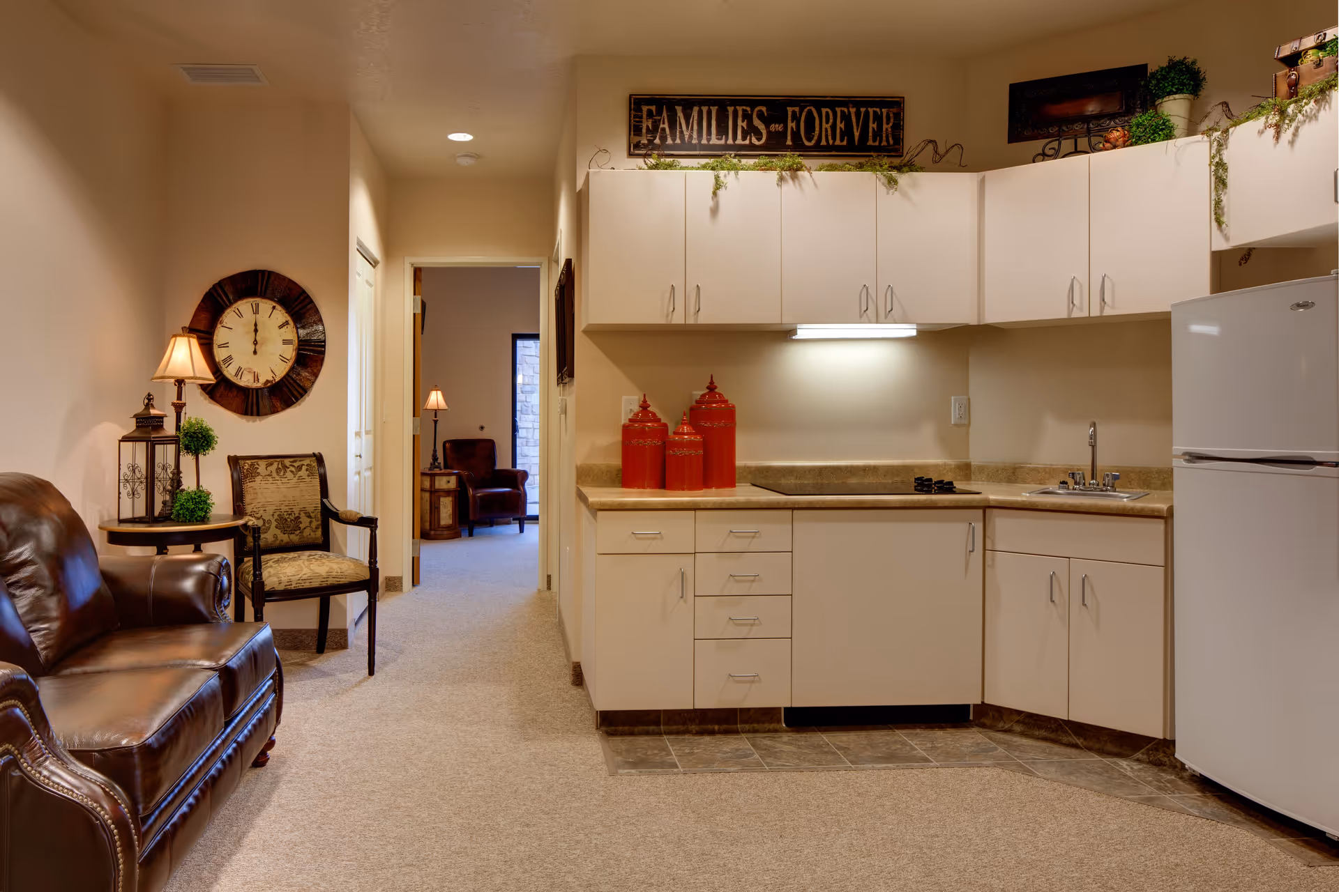 Interior view of a senior living facility room featuring a small kitchen area with white cabinets, a countertop with three red canisters, a sink, and a white refrigerator. To the left, there is a brown leather sofa, an upholstered chair, a side table with a lamp and decorative items, and a large wall clock. A hallway leads to another room with an armchair and a lamp. Above the kitchen cabinets, there is a decorative sign that reads 'FAMILIES are FOREVER' along with some greenery and decor.