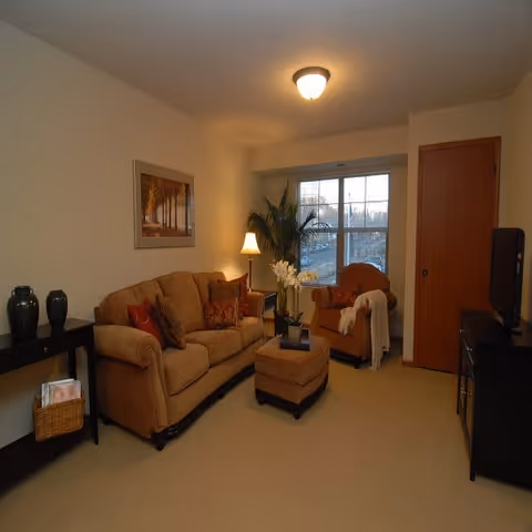 A cozy living room with a beige sofa, matching ottoman, and an armchair near a window. The room features a side table with a lamp, decorative vases on a console table, a framed picture on the wall, and a television on a stand. The room is warmly lit with a ceiling light.